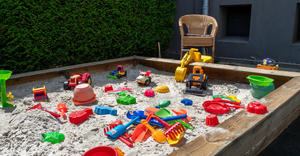 Sandkasten der Kindertagespflege Klitzeklein, gefüllt mit buntem Spielzeug und umgeben von einer Hecke.